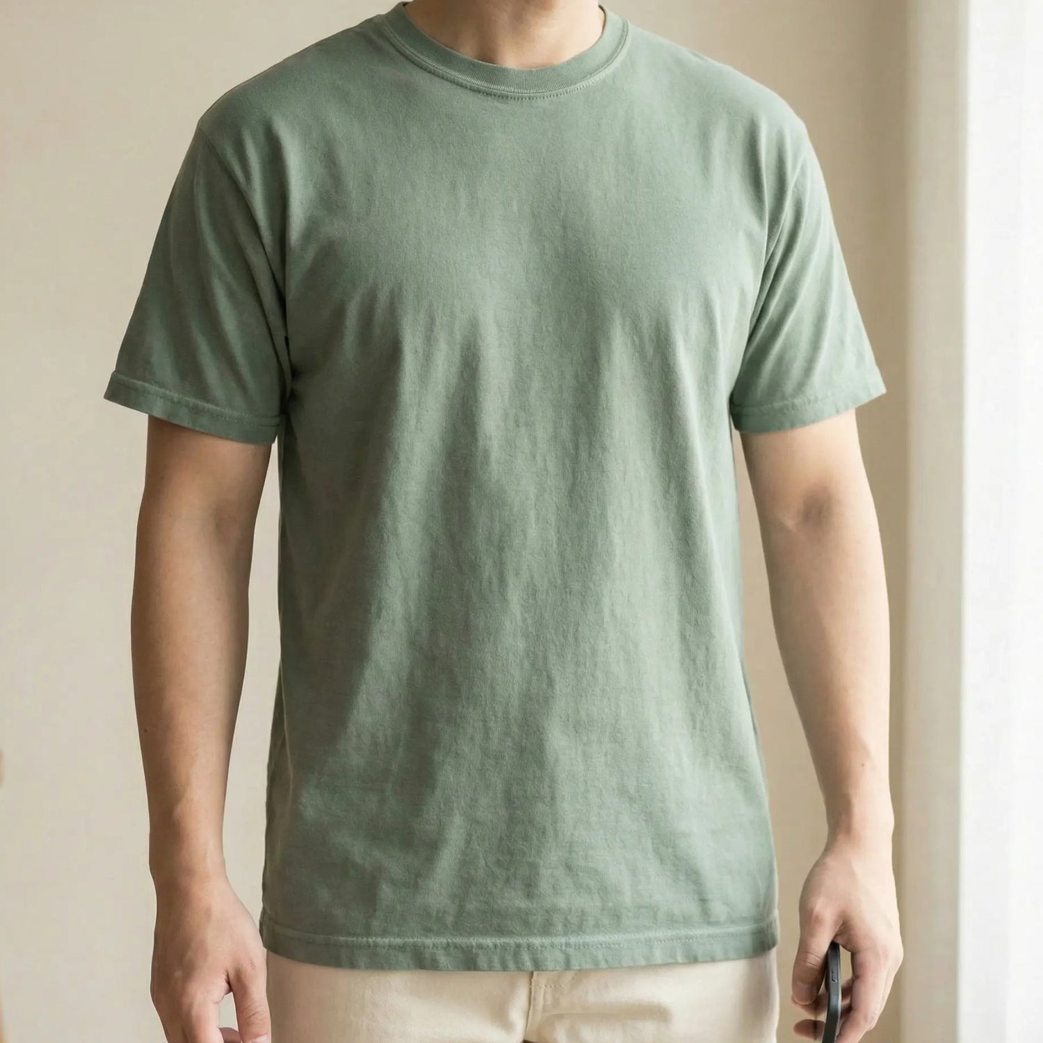 Person holding phone at side while wearing sage green minimalist t-shirt